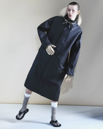 Fashion editorial shot of a slender male model in a black trench coat, socks, and sandals, with a straw hat in a studio setting.