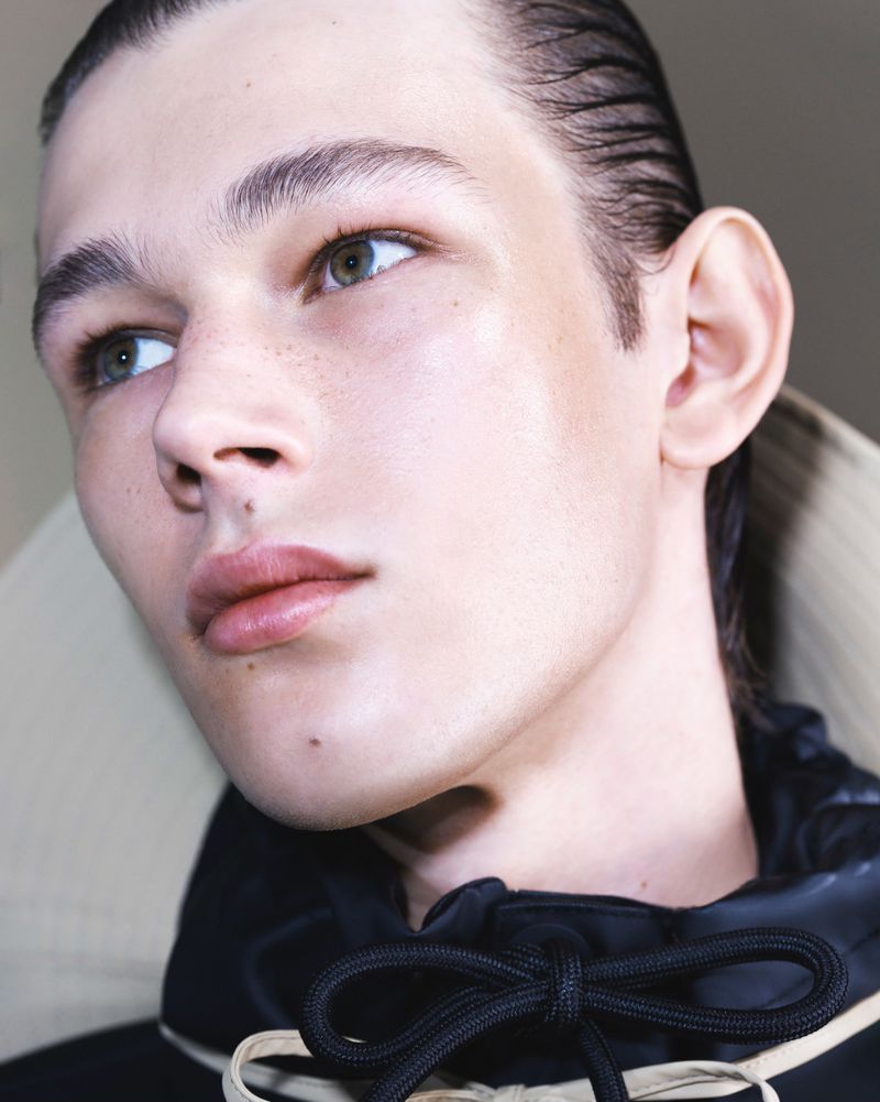 Close-up portrait of a fair-skinned young adult with slicked-back brunette hair, wearing a black satin top with tied drawstrings.