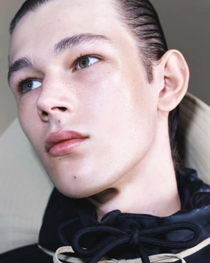 Close-up portrait of a young male model with slicked-back dark hair wearing a black jacket with drawstrings for Rains SS25.