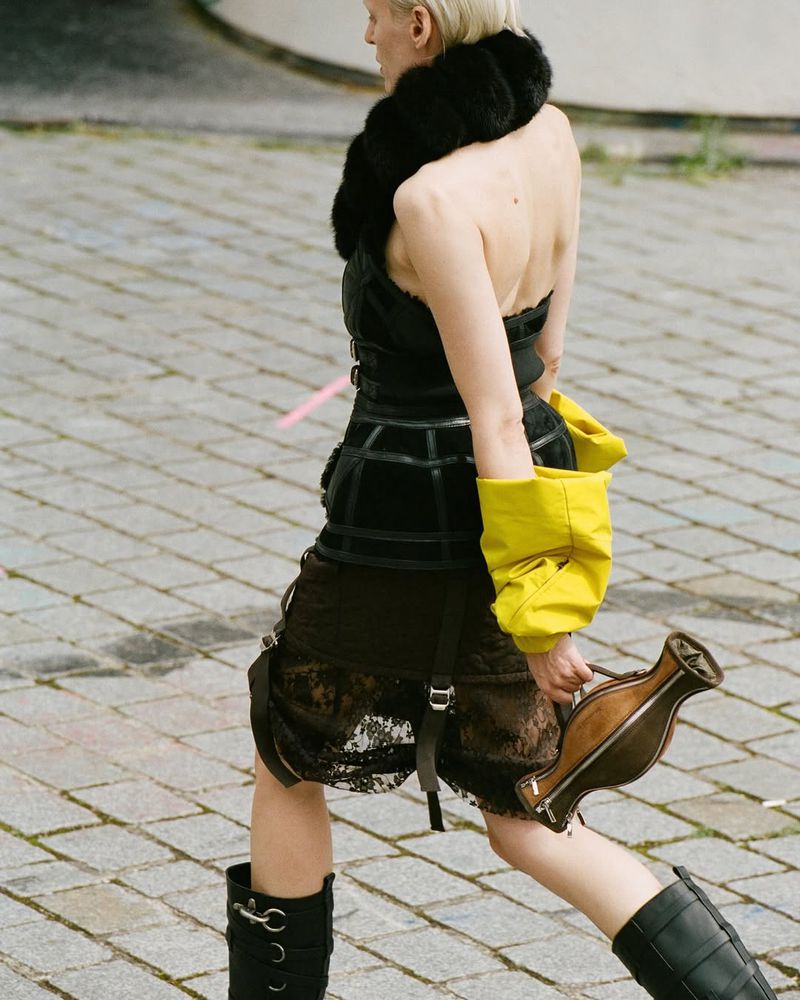 A woman models avant-garde fashion on a cobblestone street. She wears a black corset, lace skirt, black boots, and carries an unusual brown bag.