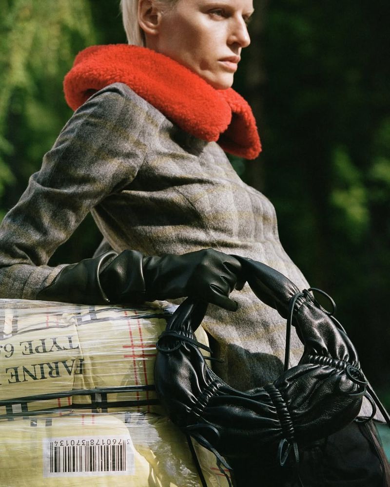 Woman in grey coat with orange collar resting on wrapped goods, holding a black bag, forest backdrop, fashion editorial, natural lighting.