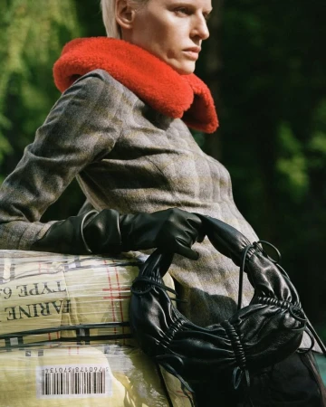 Woman in grey coat with orange collar resting on wrapped goods, holding a black bag, forest backdrop, fashion editorial, natural lighting.