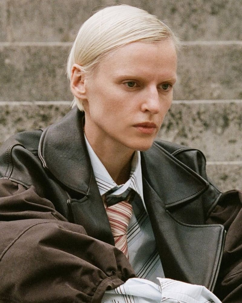 A woman with short blonde hair sits outside wearing a black leather jacket and patterned tie, captured in a medium shot.