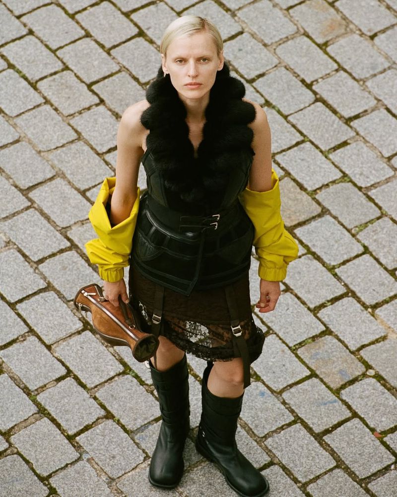 Fashion shot of a blonde adult woman standing outside on a brick surface, wearing a fur-trimmed top, yellow sleeves, skirt, and black boots.