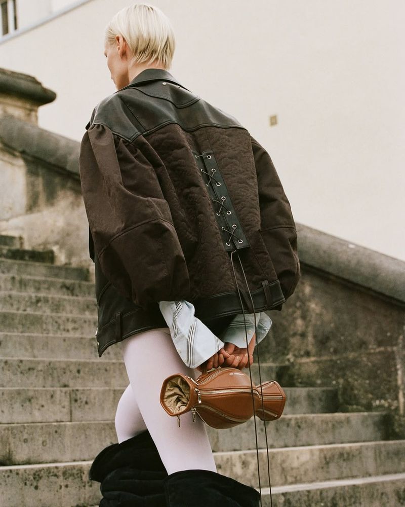 Fashionable woman with short blonde hair carries a brown leather bag on concrete stairs while wearing a brown leather jacket.