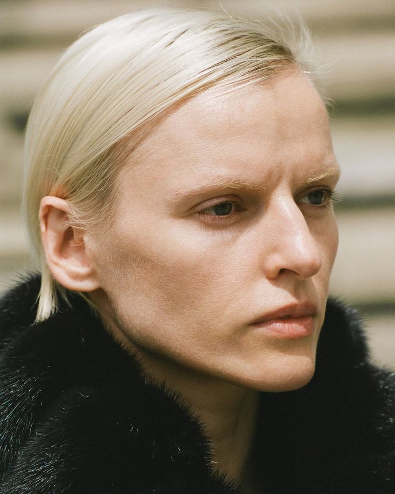 Close-up portrait of a fair-skinned adult woman with short blonde hair, wearing a dark fur coat against a blurred neutral background.
