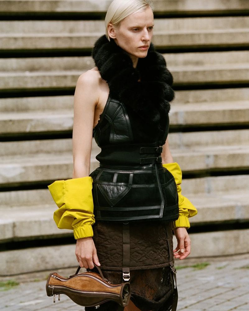 Adult female model poses in sleeveless black leather top with fur collar and yellow sleeves, holding unique purse, in front of concrete stairs.