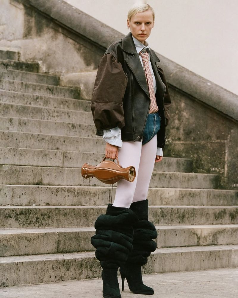 Model standing on stone outdoor stairs wearing a leather jacket, striped tie, tights, and holding a brown handbag