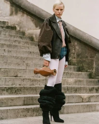 Model standing on stone outdoor stairs wearing a leather jacket, striped tie, tights, and holding a brown handbag
