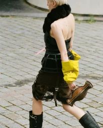 Model walking outdoors on stone paving wearing a black strapless corset top, dark skirt, and tall black boots holding a structured bag