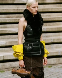 Model standing on stone steps wearing a black leather corset top, fur collar, yellow sleeves, and holding a brown bag.