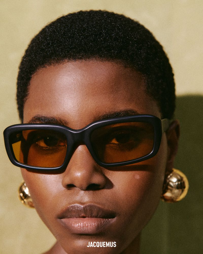 Close up portrait of a model wearing thick black sunglasses and gold sphere earrings for Jacquemus SS25