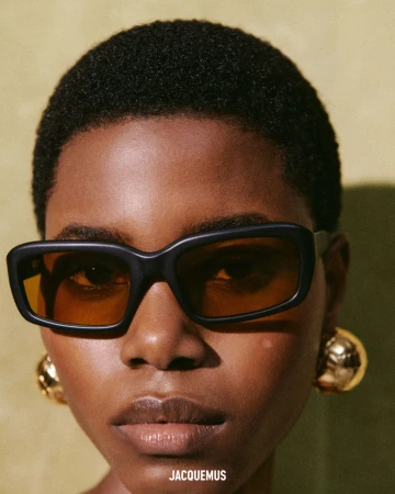 Close up portrait of a model wearing thick black sunglasses and gold sphere earrings for Jacquemus SS25
