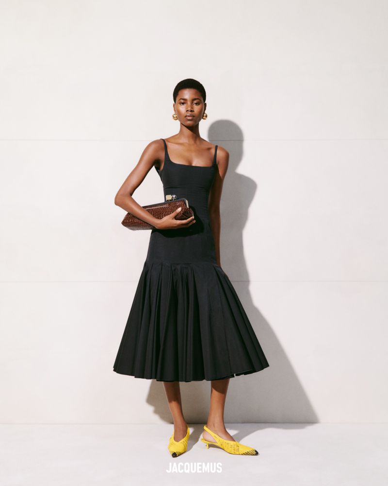 Elegant woman poses in a black dress with a pleated skirt and yellow heels while holding a brown woven handbag against a white wall.