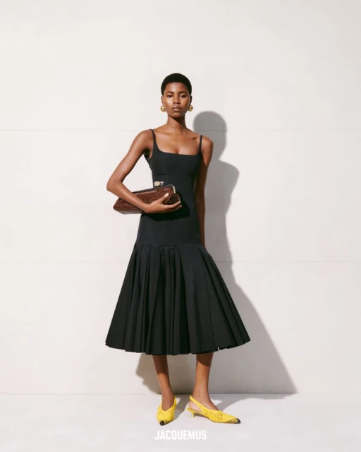 Elegant woman poses in a black dress with a pleated skirt and yellow heels while holding a brown woven handbag against a white wall.