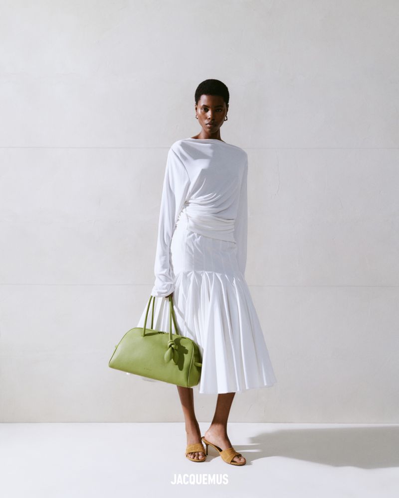 Fashion shot of an African-American woman posing in a white dress with a green handbag in a light studio against a white background.