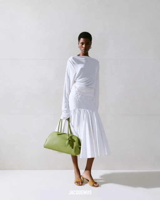 Fashion shot of an African-American woman posing in a white dress with a green handbag in a light studio against a white background.