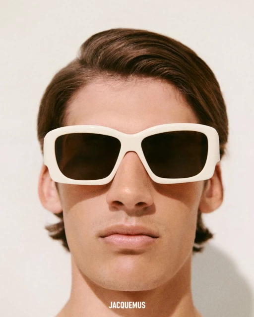Close-up studio shot of an adult male wearing square off-white sunglasses with brown hair and light skin against a white background.