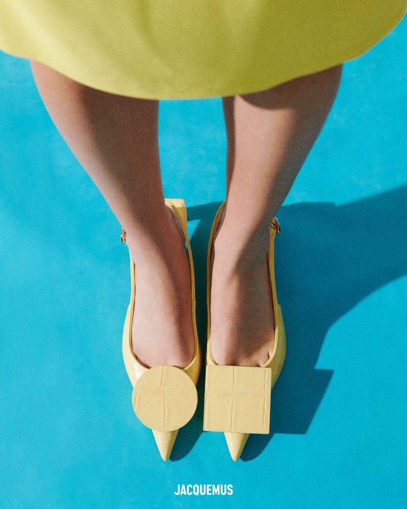 Overhead shot of a person wearing yellow heeled shoes with circle and square toe decorations on a turquoise background. Jacquemus brand.