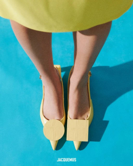 Overhead shot of a person wearing yellow heeled shoes with circle and square toe decorations on a turquoise background. Jacquemus brand.