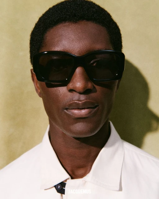 Close-up portrait of an adult Black male wearing black sunglasses and a cream-colored shirt against a solid green background.