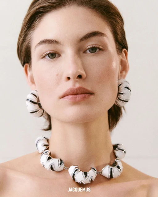Headshot of a fair-skinned woman with neutral expression wearing an art-deco style necklace and earrings with a light neutral background.
