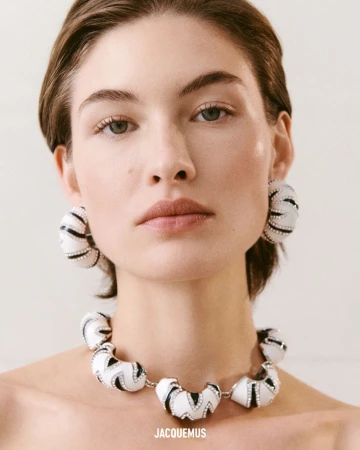 Headshot of a fair-skinned woman with neutral expression wearing an art-deco style necklace and earrings with a light neutral background.