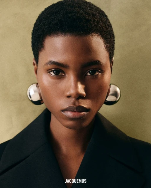 Close-up portrait of a woman with short, dark hair wearing a black blazer and large, silver ball earrings against a green backdrop.