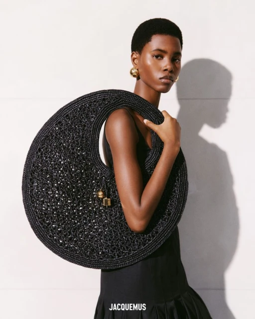 Fashion shot of an African American woman holding a black woven hobo bag, posing against a light background, wearing gold earrings.