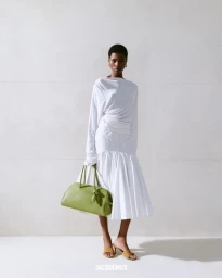 Model wearing a white dress holding a green handbag against a pale seamless studio background