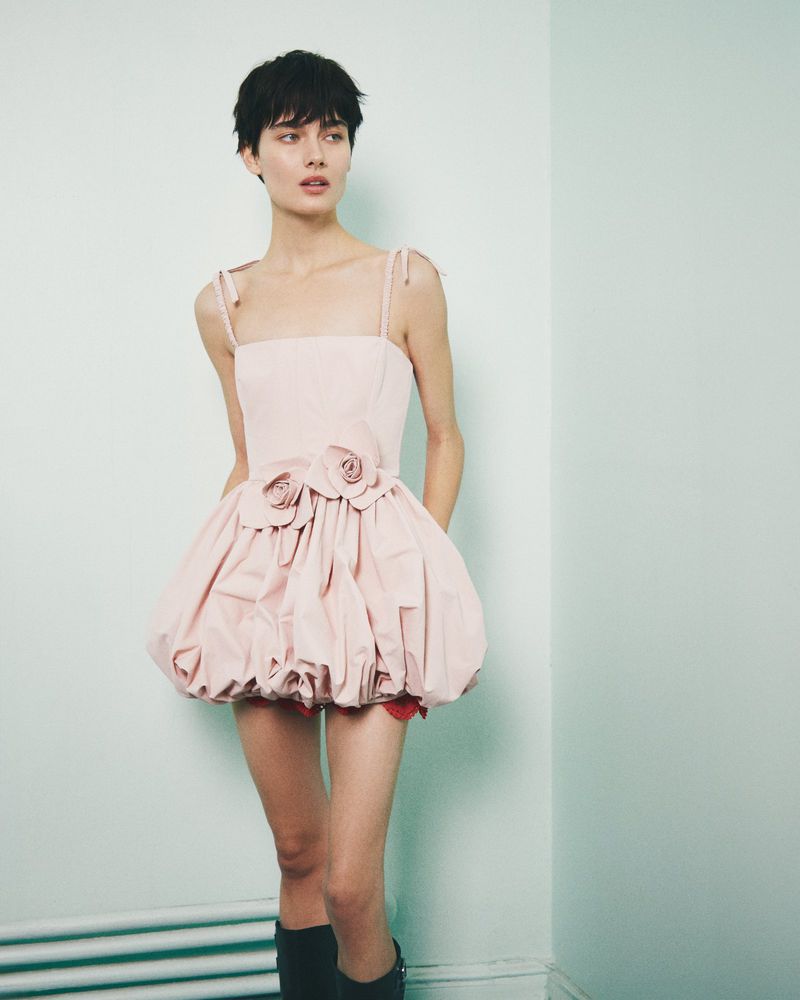 A woman with a short haircut poses in a studio wearing a pink bubble dress with two flower appliques on the bodice and black boots.
