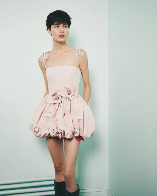 A woman with a short haircut poses in a studio wearing a pink bubble dress with two flower appliques on the bodice and black boots.