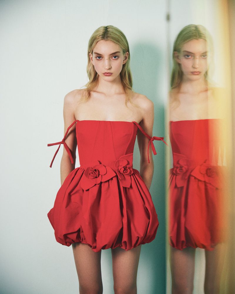 A slender female model in a red strapless dress with floral detail poses in front of a mirror in a studio setting.