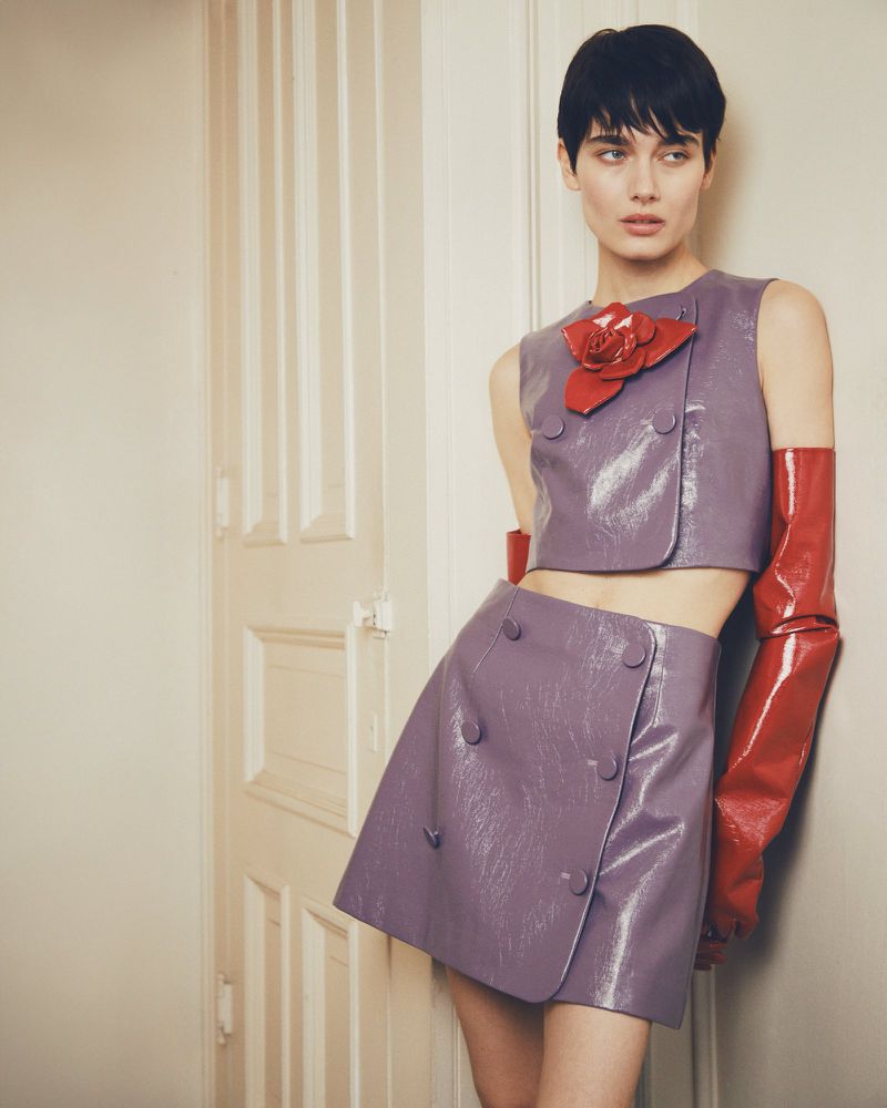 Fashion portrait of a young adult woman with short dark hair, wearing a lilac two-piece set with a red flower and gloves, leaning against a white door.