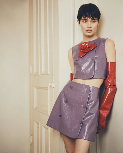 Fashion portrait of a young adult woman with short dark hair, wearing a lilac two-piece set with a red flower and gloves, leaning against a white door.