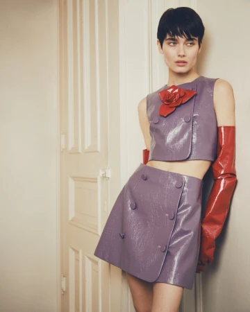 Fashion portrait of a young adult woman with short dark hair, wearing a lilac two-piece set with a red flower and gloves, leaning against a white door.