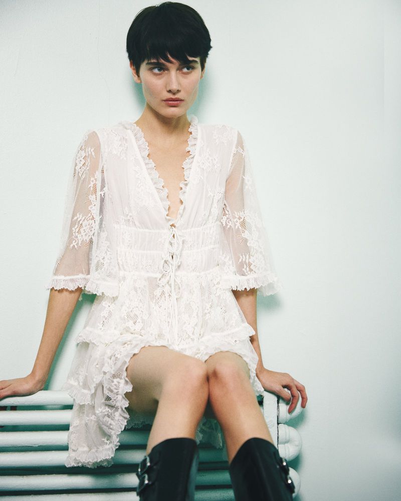 A fair-skinned woman with a pixie cut poses indoors, wearing a short white lace dress and black boots while seated on a white radiator.