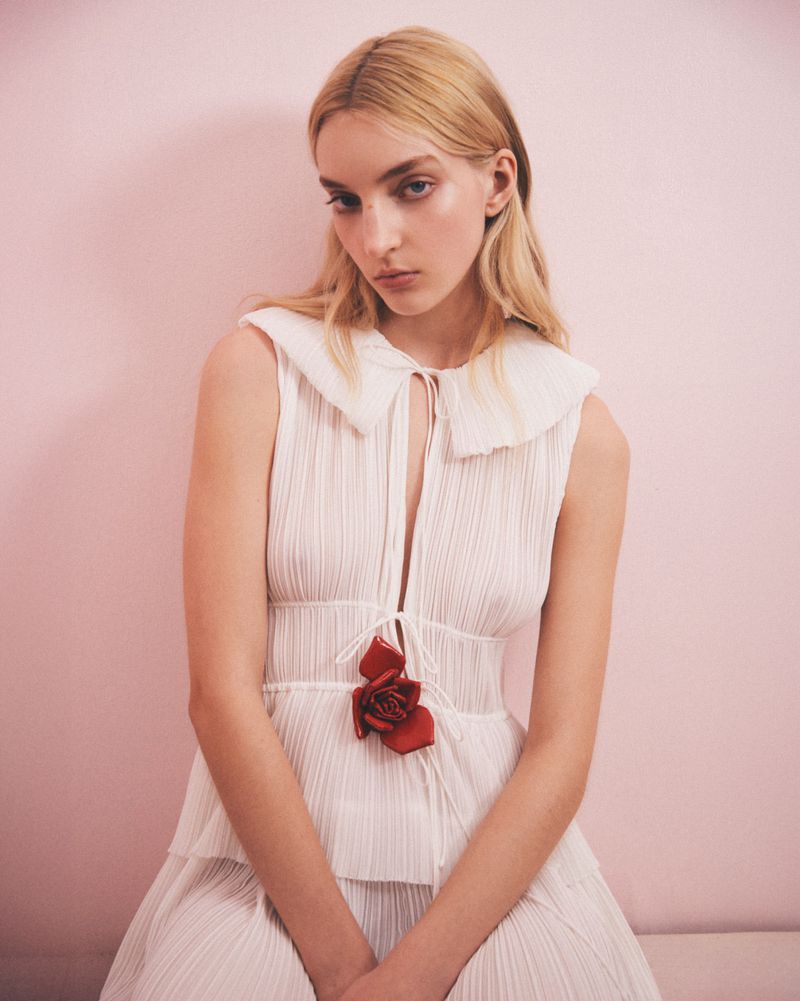 Portrait of a blonde female model in a white dress with a large collar and red rose detail against a pale pink background.