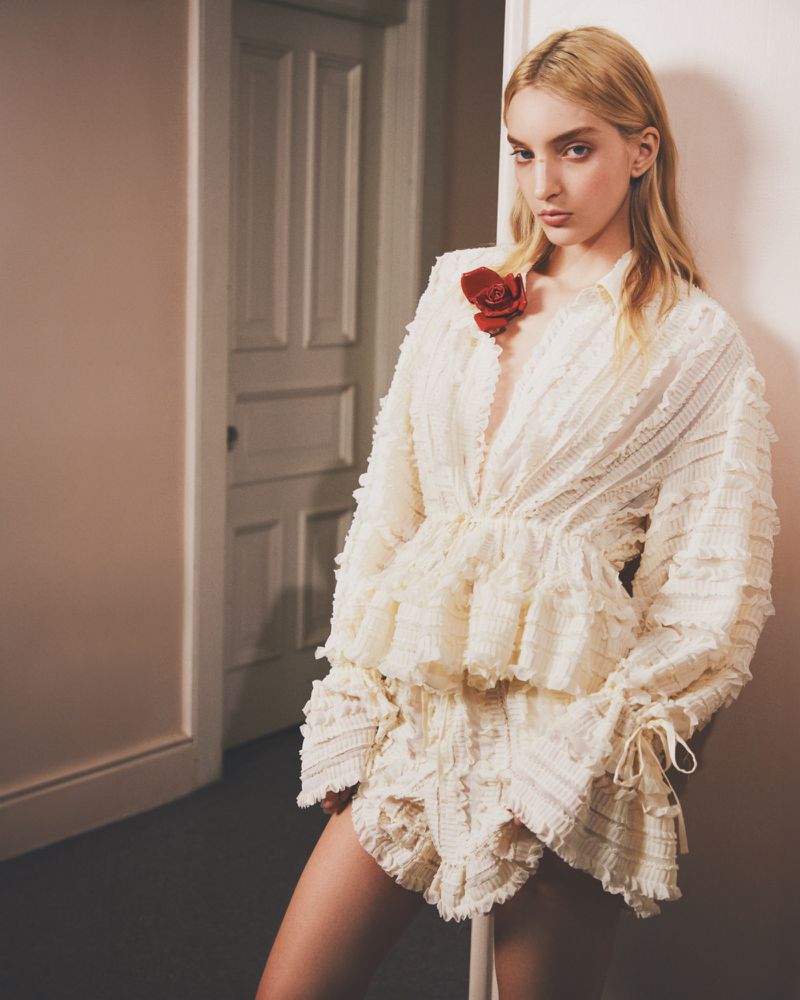 Model in ruffled white blouse with red flower pin leaning against a wall in a domestic interior for Aknvas