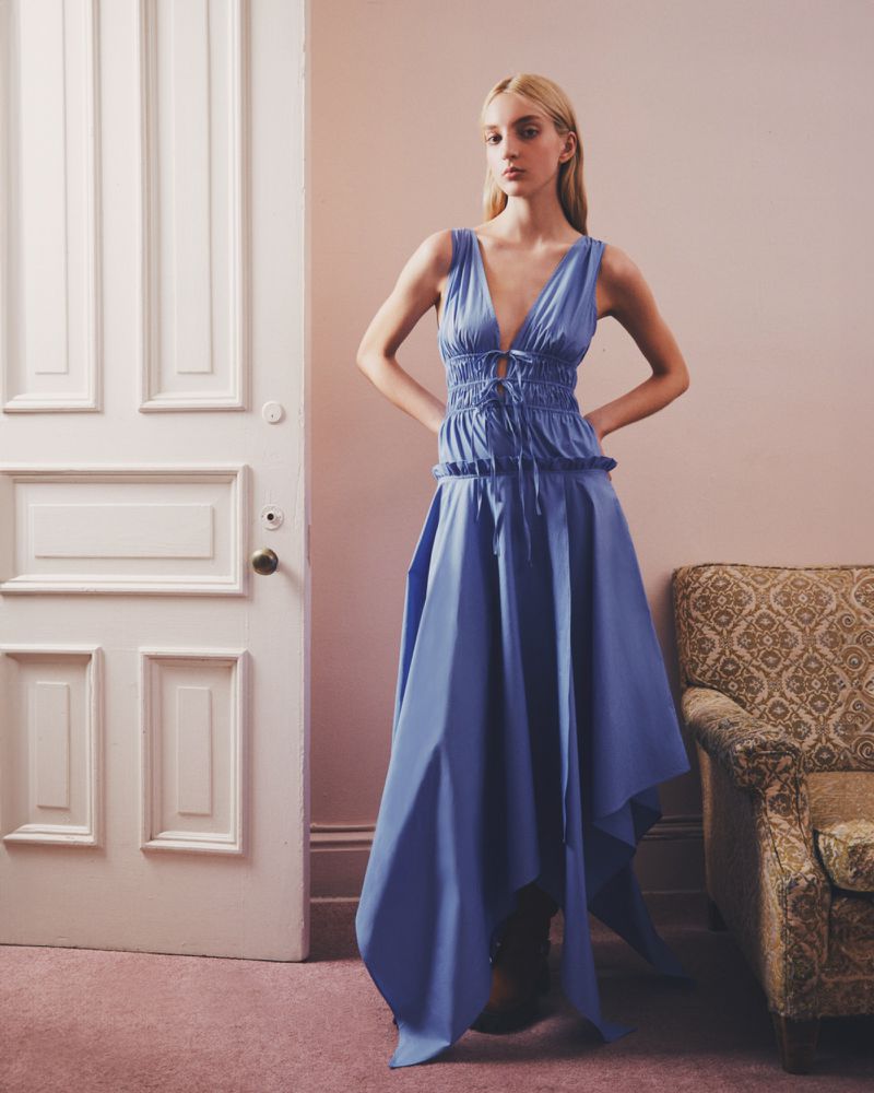 A fair-skinned woman poses indoors wearing a long, periwinkle-blue dress in a fashion editorial, with a chair and door in the background.