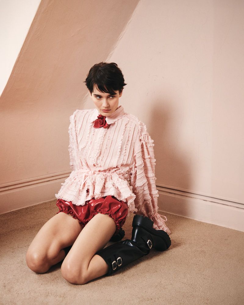 Woman in frilly pink blouse and red shorts kneeling on floor with black boots in pink room with natural light casting soft shadows.
