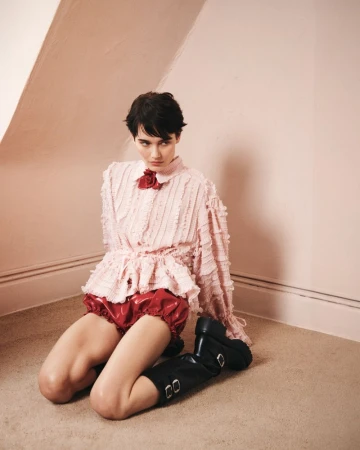 Woman in frilly pink blouse and red shorts kneeling on floor with black boots in pink room with natural light casting soft shadows.
