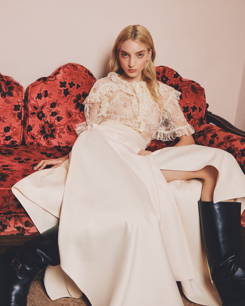 A woman poses on a red floral couch, wearing a white lace top, a long white skirt and tall black boots in an indoor setting.