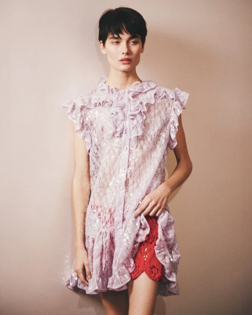 A model with short dark hair poses in a lilac lace dress with a ruffled collar and a red lace slip showing underneath. Fashion editorial shot.