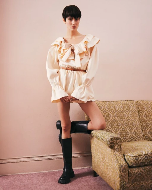 Woman in ruffle-collar romper and knee-high boots poses on a couch in a studio with a neutral background.