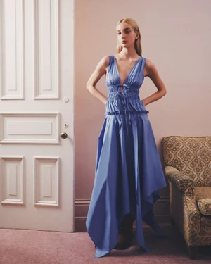 Model standing in a blue sleeveless V-neck dress inside a room with a white paneled door and patterned armchair.