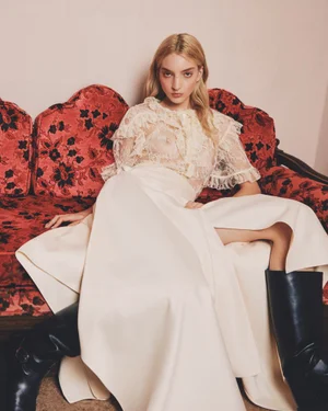 Model seated on a red patterned vintage sofa wearing a sheer lace blouse and cream skirt with black leather boots.