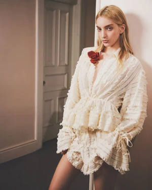 Model in ruffled white blouse with red flower pin leaning against a wall in a domestic interior for Aknvas
