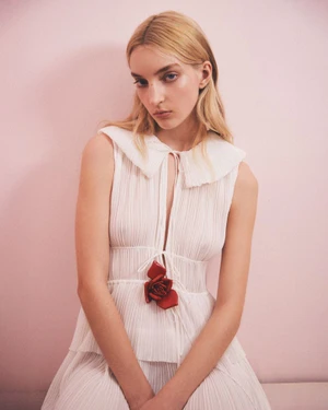 Blonde female model wearing a white pleated dress with a red rose brooch against a pale pink background for Aknvas Pre-Fall 2025 Lookbook