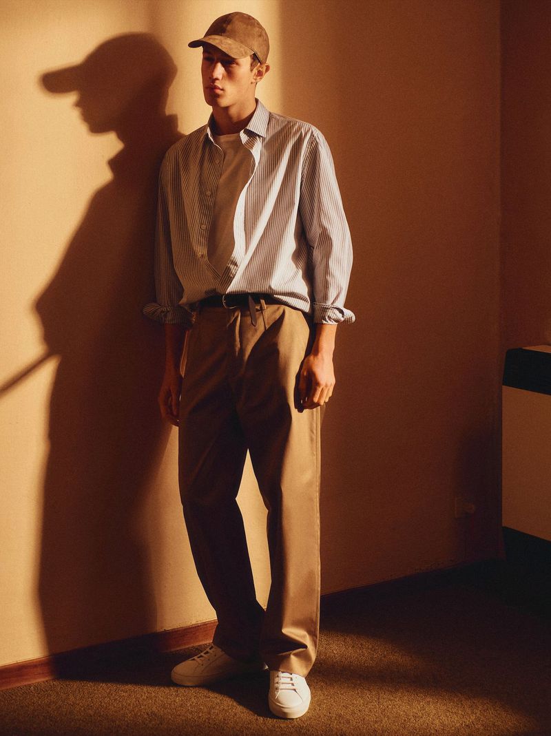 Young adult man in casual outfit posing indoors, leaning against a wall. He is wearing a shirt, trousers, cap, and white sneakers.